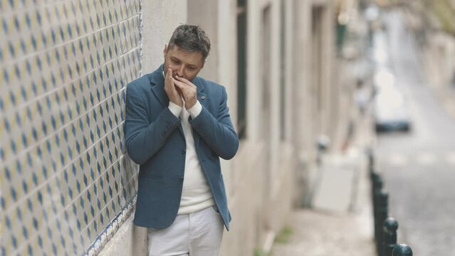A man in a blue jacket stands against the wall and plays the harmonica in the street