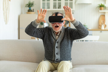 Excited mature senior man wearing using virtual reality metaverse VR glasses headset at home....