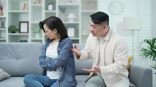 Angry asian husband arguing with wife sitting on sofa in living room at home. Conflict and dispute between a married couple. A man quarrels with a sad woman. Problem and crisis in family relations