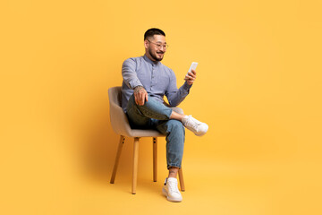 Fototapeta na wymiar Young Asian Man Using Smartphone While Sitting In Chair Over Yellow Background