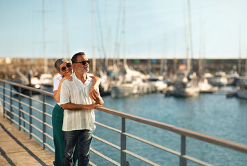 Senior Couple Hugging At Marina Pier Looking At Yachts Outdoors