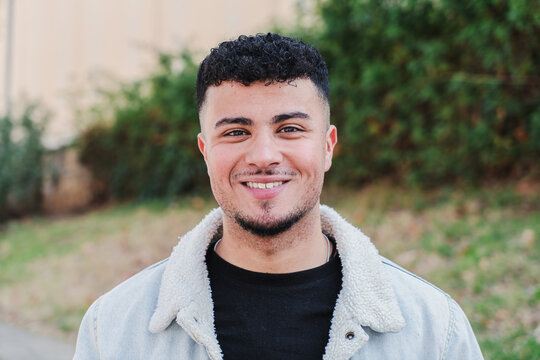 Close up portrait of happy young man smiling and looking at camera. Front view of male teenager standing outdoors at university campus. Student guy laughing at high school. Youngster person face. High