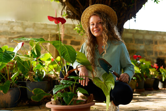 Young woman gardener in straw hat holding hand shovel taking care of potted plants. Junior caucasian female smiling sitting in her little garden planting flowers in pots. Gardening and farming concept