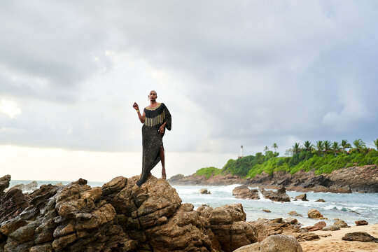 Non-binary black godlike person poses gracefully standing on rocks in ocean. Trans ethnic fashion model in posh dress and jewellery on rocky beach by storm . Lgbtq. Pride month