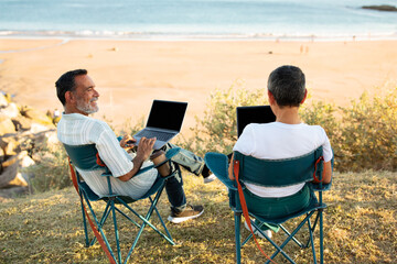 Senior Couple Using Laptops Sitting At Beach Near Ocean Outdoor