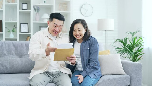 Happy asian couple celebrating success reading a letter with great news sitting on sofa in room at home. Excited smiling joyful husband and wife hugging, rejoicing, pleased with pleasant notification