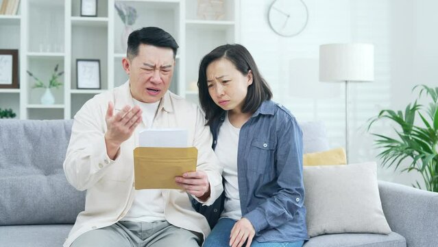 Disappointed asian couple reading letter with bad news sitting on sofa in room at home. Sad husband and wife are depressed by an unpleasant notification, shocked by information about failure, losses