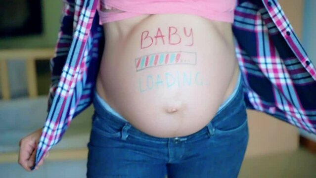 Blue and pink baby loading inscription on woman's pregnant belly, expecting mom waiting for her baby arrival, happy female slowly dancing and posing on camera, tenderness and motherhood concept