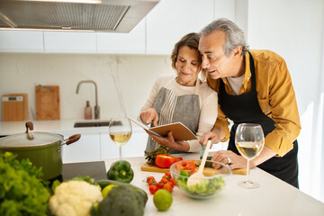 Cooking together. Senior spouses reading recipe in cookbook while preparing lunch together in kitchen, copy space