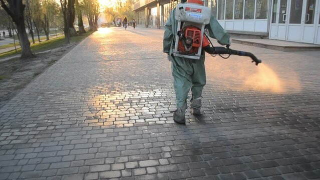 Man in airtight suit sprays disinfecting liquid a pavement on a street in the city against the backdrop of a sunset. Sanitation workers clean a crosswalk. Sanitary measures. coronavirus pandemic
