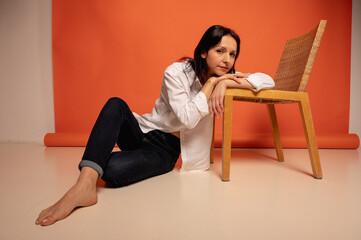 Full length photo of attractive chic businesswoman sitting on floor leaning on chair, flirtatious mood, blank space in jeans and white shirt on orange background, place for text