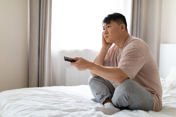 Unhappy middle aged asian man sitting on bed, watching TV