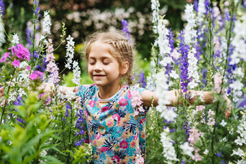 A funny little girl has fun in the summer outdoors. A carefree child runs and jumps among blooming summer flowers. Portrait of a beautiful girl smiling contentedly while smelling flowers in the garden