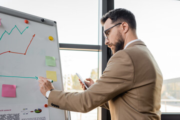 side view of accomplished businessman in beige suit and eyeglasses holding mobile phone and drawing on flip chart while making productivity analysis in office