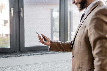partial view of bearded businessman in beige stylish blazer holding mobile phone with blank screen while standing near large windows in contemporary office