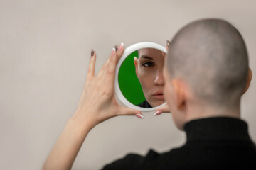 a young slender woman with a shaved bald head stands with her back to us and examines herself in the mirror, selective focus. Side effects of chemotherapy during cancer treatment