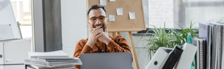 overjoyed businessman in trendy eyeglasses covering mouth with hands and laughing near laptop,...