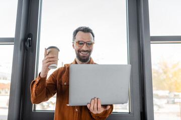 successful bearded businessman in trendy shirt and eyeglasses holding coffee to go and pointing up with finger during video call on laptop in modern office, corporate lifestyle