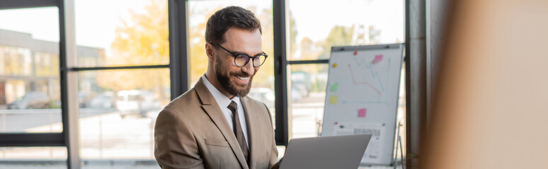 joyful bearded manager in eyeglasses, beige blazer and tie looking at laptop during video...