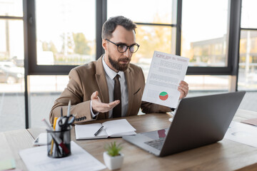 Fototapeta na wymiar bearded businessman in eyeglasses, tie and beige blazer showing document with market research and pointing with hand during video conference on laptop near notebook, smartphone and office stationery