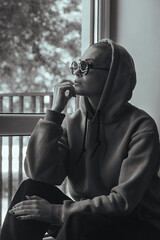 A beautiful young woman with a shaved head without hair sits and looks out the window, she is wearing dark glasses and a hoodie. Black and white photo