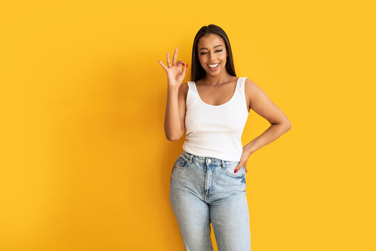 Young woman wearing casual clothes, white top and jeans,  showing okay sign with fingers, smiling to the camera. Yellow studio background with copy space