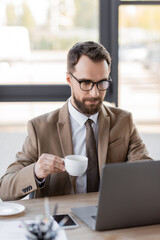Fototapeta na wymiar focused bearded businessman in stylish blazer, eyeglasses and tie holding coffee cup near mobile phone with blank screen and working on blurred laptop in office