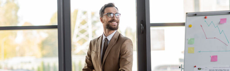 Fototapeta premium successful bearded businessman in stylish eyeglasses, beige blazer and tie looking away and smiling near flip chart with graphs and sticky notes in office with large windows, banner