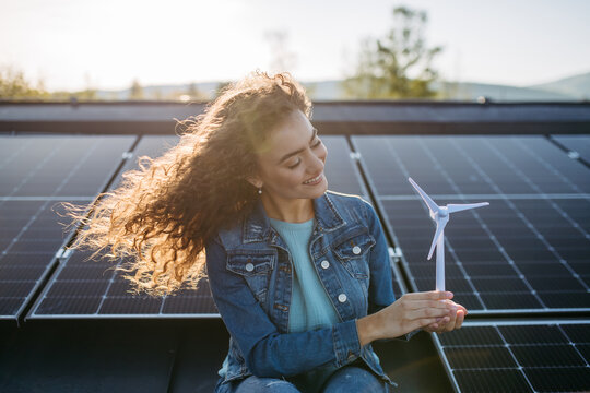 Portrait of young woman holding model of wind turbine on roof with solar panels.