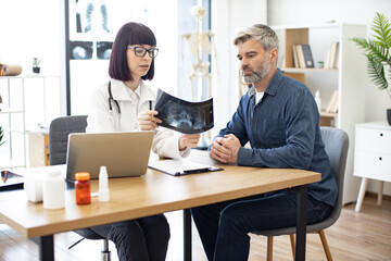 Skilled radiologist in white uniform and eyeglasses explaining visible changes on x-ray image. Anxious man dressed in jeans and denim shirt carefully listening to physician's instructions.
