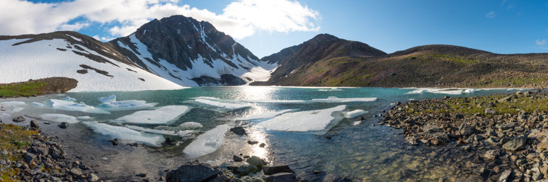 Panoramic shot of glacial lake in northern Canada during summer time on beautiful blue sky day. Large ice chunks in water, creek, river. Paddy Peak, Yukon Territory. 