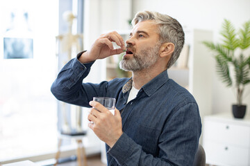 Caucasian middle-aged caucasian man taking pill and drinking glass of fresh water in doctor's office. Grey-bearded male person dressed in denim shirt following cardiologist's instructions. - obrazy, fototapety, plakaty