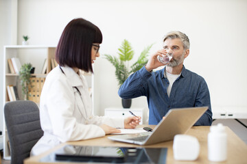 Fototapeta premium Grey-haired patient drinking glass of cold water at appointment with general practitioner in new clinic. Young professional writing out prescription and using remote computer for working with clients.