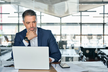 Serious mature business man executive sitting at desk using laptop. Busy professional mid aged...