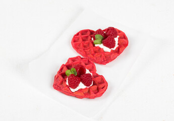 Valentine's Day dessert. Two red heart-shaped waffles with cream, raspberries and micro greenery. On serving paper. White background