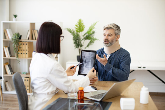 Mature bearded man in C collar talking to female physician in white coat pointing at tablet with spine scan on screen. Worried patient explaining health condition to medical person in consulting room.