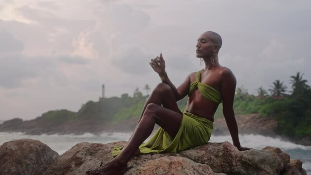 Queer black person poses in open dress, handmade jewelry sits at stone with scenic sea cliff top above dramatic cloudy sky, ocean waves. Lgbtq biethnic gay model in brass accessories. Tropical sunset.