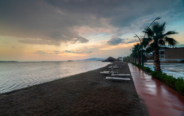 sunbeds tables and sunshade umbrellas located by the sea, trees and beach sand with a colorful sunset after the rain