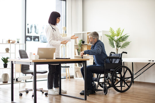 Young woman in lab coat helping mature man with walking cane to stand up from wheelchair in consulting room. Efficient therapist assisting patient in finding balance during recovery from injury.