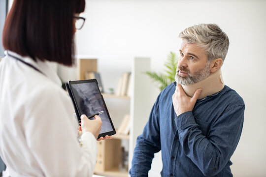 Caucasian lady holding tablet with CT scan image on screen while talking to male in neck brace sitting on exam couch. Young orthopedist analyzing thoracic vertebrae on digital report in clinic. - Powered by Adobe