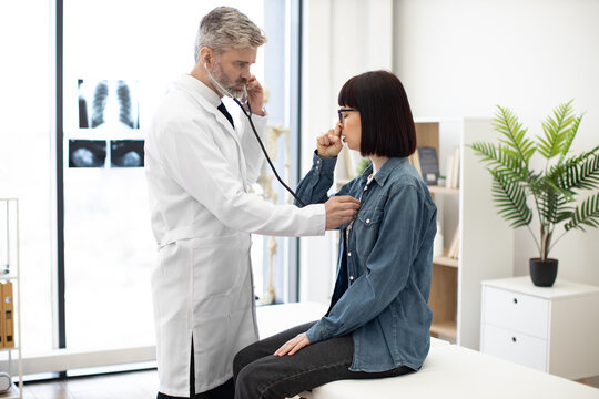 Beautiful woman in jeans outfit coughing while sitting on exam couch during medical checkup in private practice. Focused family doctor listening to lungs of lady with stethoscope in consulting room.