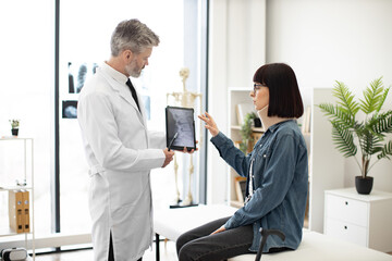 Confident mature man in lab coat holding digital tablet while young lady in cervical collar sitting...