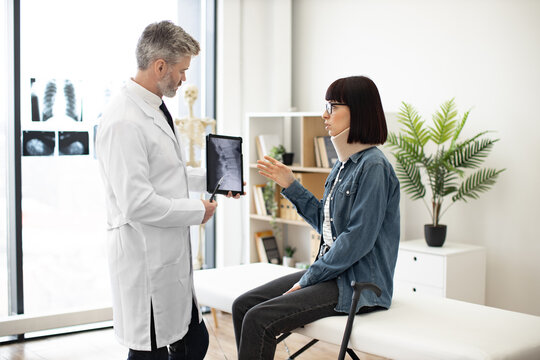 Caucasian woman in neck brace looking at x-ray image on digital device held by bearded therapist in lab coat. Medical specialist explaining test results to patient with walking cane in exam room.
