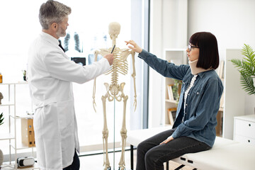 Side view of mature man in doctor's coat pointing at skeleton while young woman in glasses and neck...