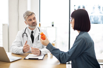 Side view of pretty woman wearing jeans outfit looking at medications in glass container in clnic. Caucasian female in glasses questioning aged male doctor about side effects of pills.