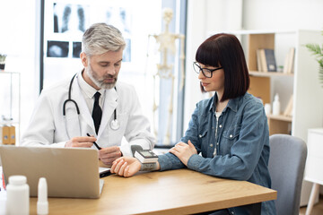 Confident bearded male with stethoscope turning on wearable tonometer applied on female wrist at doctor's office. Focused aged family physician measuring pulse rate of young caucasian woman.