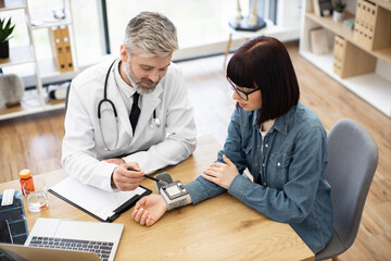Obraz premium Top view of bearded adult with stethoscope and brunette in denim outfit monitoring electronic tonometer on wrist. Cheerful physician analyzing positive results tracked by device at doctor's office.