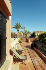 Outdoor terrace with earth tone colors, plants, palm trees, resting chairs and a blue sky on a beautiful sunny day in a riyad hospitality house in Marrakech, Morocco