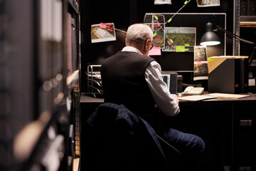 Elderly fbi investigator sitting at desk table in arhive room, analyzing crime evidence files....