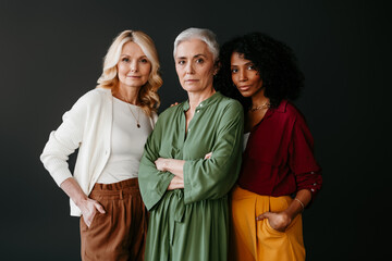 Three confident mature women looking at camera while bonding against grey background
